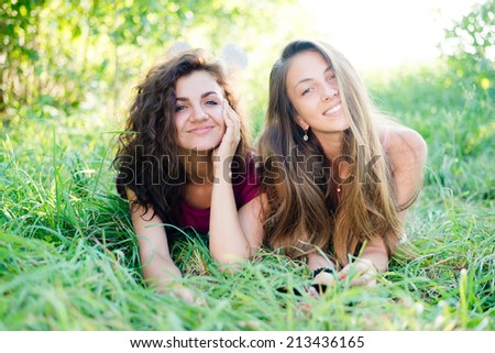 2 playful pretty girlfriends having fun laying in grass happy smiling & looking at camera on green summer outdoors copy space background, portrait