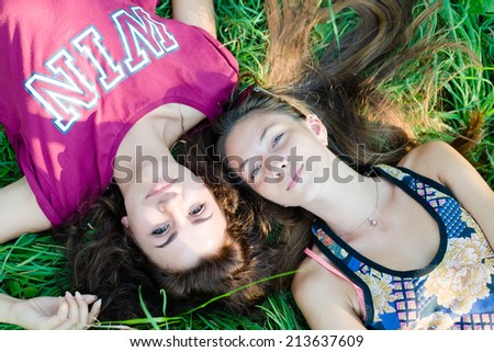 portrait of 2 beautiful brunette young women best friends having fun relaxing & happy smiling looking at camera while lying on green grass outdoors copy space background