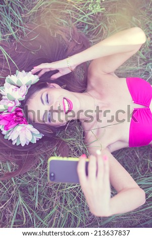 fairy selfy: filtered image of beautiful brunette young lady in pink bikini and flower crown having fun making selfie picture lying on green grass outdoors copy space background