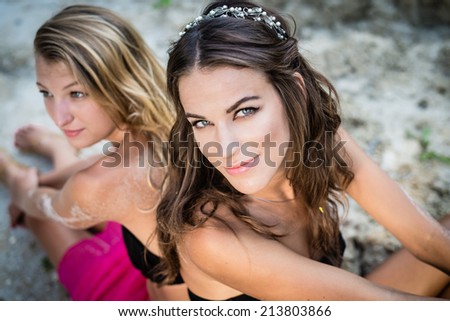 2 beautiful brunette and blonde best girlfriends wearing silver diadems sitting back to back with bare shoulders on sandy cracked ground outdoors background, closeup portrait