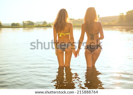 filtered image of 2 attractive young women or teenage girls best friends in bikini holding hands and looking at summer river or lake on outdoors copy space background