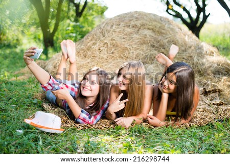 closeup portrait of 3 pretty girls having fun relaxing lying on hay happy smiling with excellent white teeth making selfie & looking at mobile phone on green summer outdoors copy space background