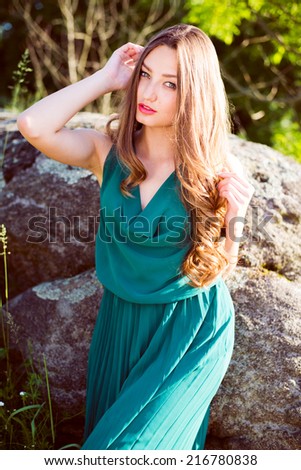 sensual beauty: young beautiful woman sitting on stone in green dress with sun light flares of rays & looking at camera on summer outdoor copy space background