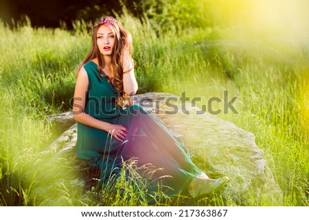 portrait of brunette young beautiful woman sitting on stone in green dress with pink wreath of flowers & sun light flares of rays & looking at camera on summer outdoor copy space background