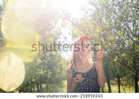 pretty apple fairy: young woman cute blond girl smiling among red apples with wreath of flowers & sun light flares of rays on summer outdoor copy space background