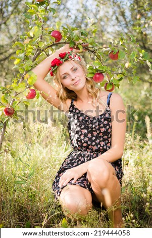 pretty apple fairy: young woman cute blond girl smiling among red apples with wreath of flowers & sun light flares of rays looking at camera on summer outdoor copy space background