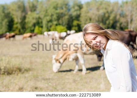 joyful cowgirl: active beautiful young woman in white shirt having fun and smiling among cows looking to camera on green forest copy space background