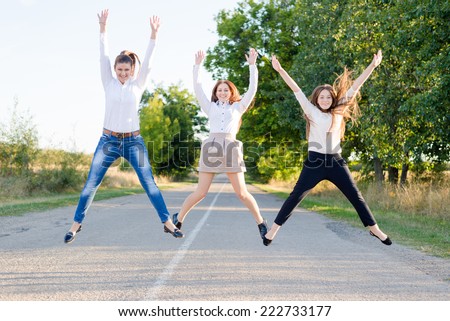 Back to school: three active beautiful young female students having fun jumping high on country road looking to camera on green trees copy space background