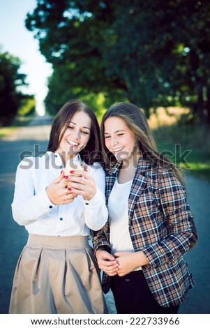 Filtered portrait of 2 happy pretty sisters in formal wear looking on mobile phone and laughing on green outdoors copy space background