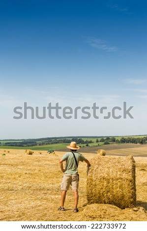 picture of man standing in the golden field of rounded hay stock. farmer standing on summer blue sky copy space background open horizon.