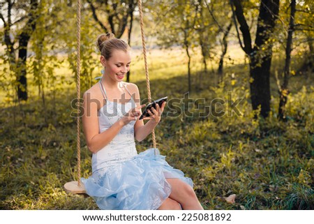 Beautiful young woman in prom dress sitting on swings and typing on tablet pc on green summer or autumn outdoors copyspace background
