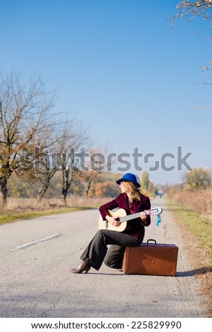 Young retro woman musician in retro hat sitting on vintage suitcase and playing guitar on empty autumn road copy space background