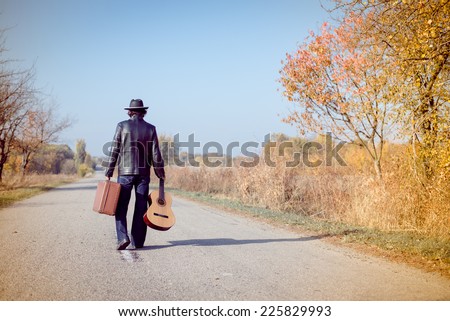 Young man musician in retro hat and leather jacket with vintage suitcase and guitar walking away on empty autumn road copy space background