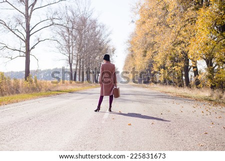 Young lady in retro hat and overcoat with vintage suitcase waiting on empty autumn road copy space background