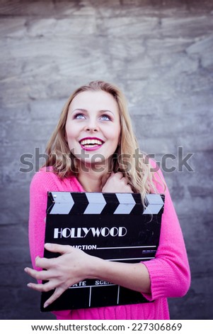 Happy young blond woman holding cinema clapper board and smiling over gray concrete or brick wall copy space background
