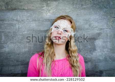 Young blond sensual woman in white lace mask over gray brick wall copy space background