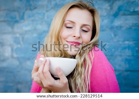 Happy young blond woman enjoying hot tea or coffee and smiling over blue brick wall copy space background