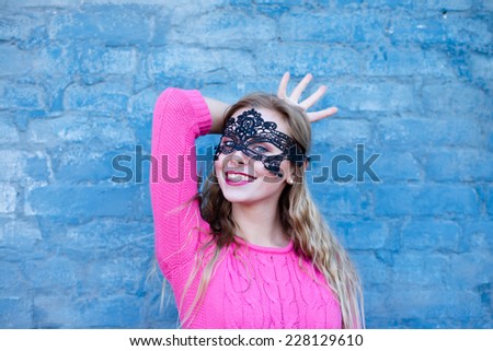 Happy young blond woman in black lace mask pretending to be queen with four fingers as crown over gray brick wall copy space background