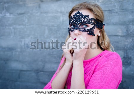 Young blond woman in black lace mask looking scared over gray brick wall copy space background