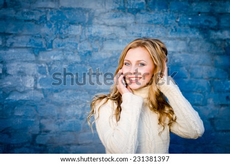 image of beautiful blond young lady in white wool sweater speaking on mobile at blue concrete wall copy space background