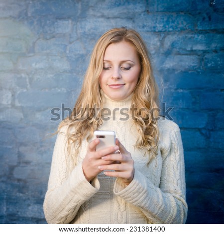image of beautiful blond young lady in white wool sweater texting mobile message at blue concrete wall copy space background