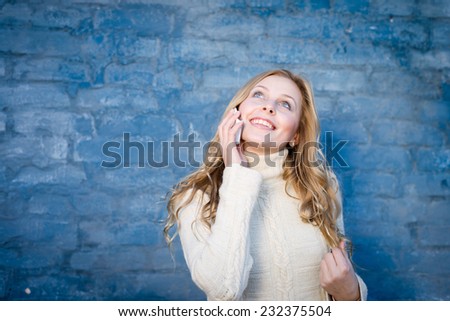 image of beautiful blond young lady in white wool sweater speaking on mobile at blue concrete wall copy space background