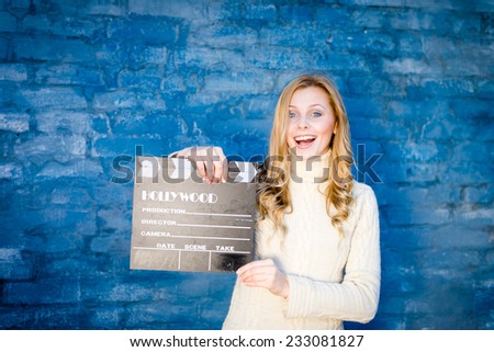 Happy young blond woman in white knitted sweater with cinema clapper board smiling over blue concrete or brick wall copy space background