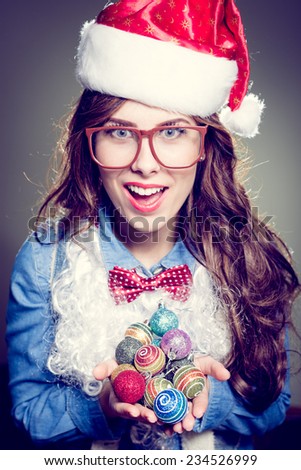 Funny hipster girl in super size eyeglasses wearing xmas santa hat and faux beard holding pile of Christmas tree decoration balls over olive copy space background