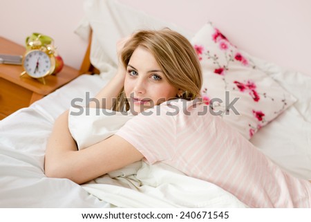 portrait of beautiful happy young blond lady in bed with alarm clock & floral pillow in the background