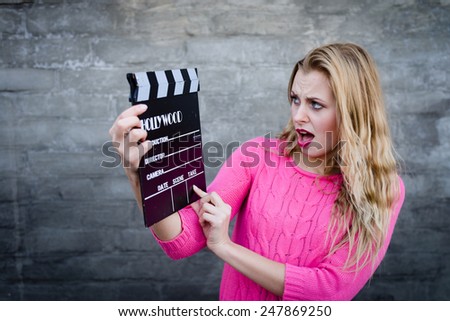 Happy young blond woman in pink knitted sweater with cinema clapper board looking surprised over brick wall copy space background