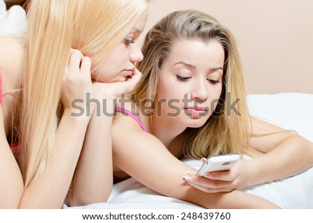 looking at mobile cell phone 2 adorable blond sisters or beautiful sexy girl friends having fun relaxing on a white bed