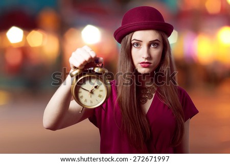 Portrait of showing alarm clock elegant beautiful young woman in hat looking in camera over bright party bokeh background