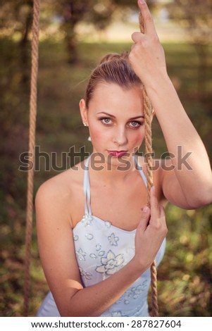 Beautiful young woman in prom dress sitting on swing on green summer outdoors