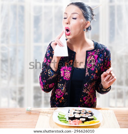Young brunette pretty woman touching her mouth with table napkin standing in front of table with plate of sushi and happy smiling