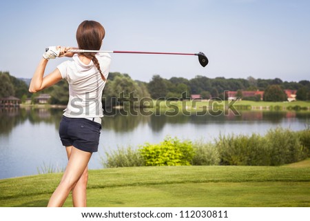 Girl golf player teeing-off from tee-box with driver, view from behind.