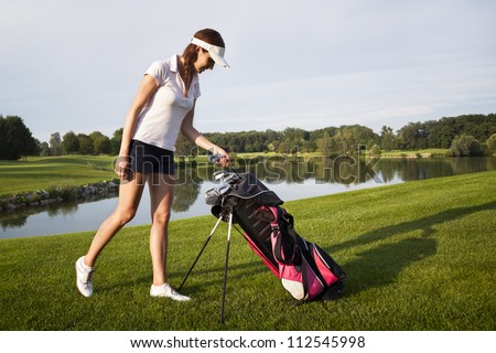 Woman golf player taking out golf club from golf bag on golf course with beautiful pond and trees in background.