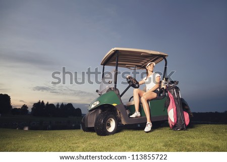 Smiling woman golf player sitting in golf cart at sunset with golf bag and clubs.
