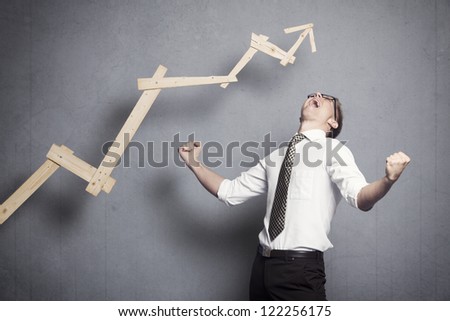 Concept: Great achievement in business. Enthusiastic young businessman cheering in front of business graph with positive trend, isolated on grey background.