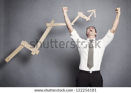 Concept: Success in business. Overjoyed talented businessman with raised arms cheering in front of positive business graph, isolated on grey background.