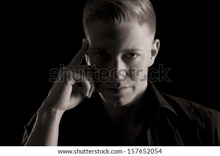 Low-key close up portrait of young serious man in dark shirt looking straight, black and white, isolated on black background.