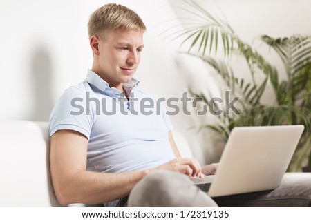 Young smiling man sitting relaxed at home on sofa working on laptop.