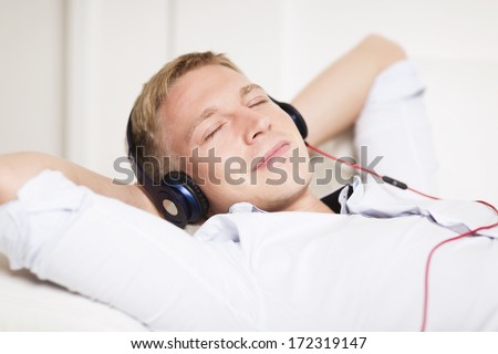 Close up portrait of young man relaxing with hands behind head and listening music at earphones while lying down on couch.
