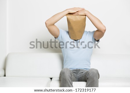 Desperate anonymous man with head covered by a blank paper bag sitting on sofa with hands on head and looking down, empty space for text, on white background.