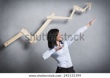 Concept: Successful business trend. Happy talented businesswoman pointing arm upwards in front of ascending business graph, isolated on grey background.
