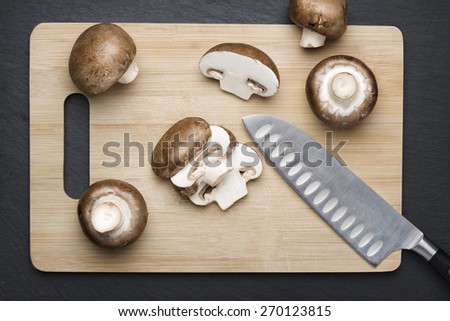 Several brown sliced champignon mushrooms on a wooden chopping board with knife, with dark grey slate stone background.