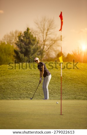 Woman golf player putting on green looking at flag, with sunset in background.