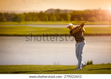 Male senior golf player swinging golf club with lake in background at sunset with copyspace.