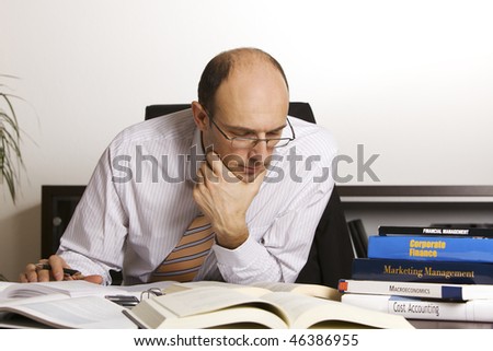 Businessman in suit sitting at desk in office in front of a stack of business books studying information, white background