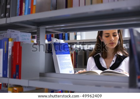 View through bookshelf at attractive student standing in between bookshelves in modern university library reading a book.