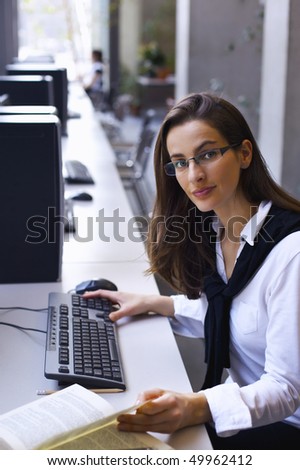 Young attractive woman sitting at PC in computer lab working and  studying a book.